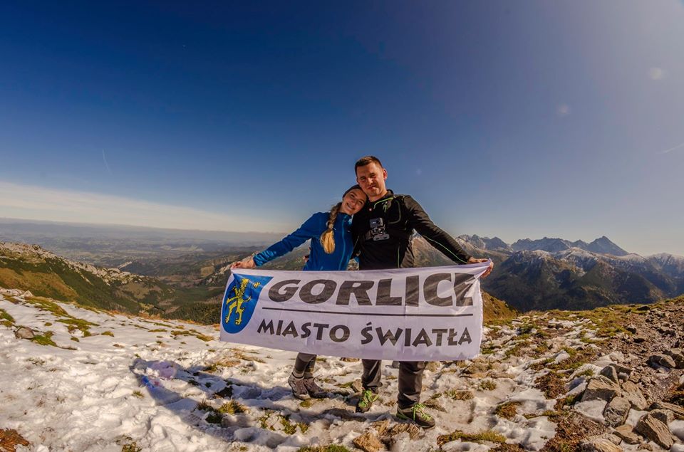 Dwie osoby stoją na szczycie z flagą Gorlice Miasto Światła w ręku.